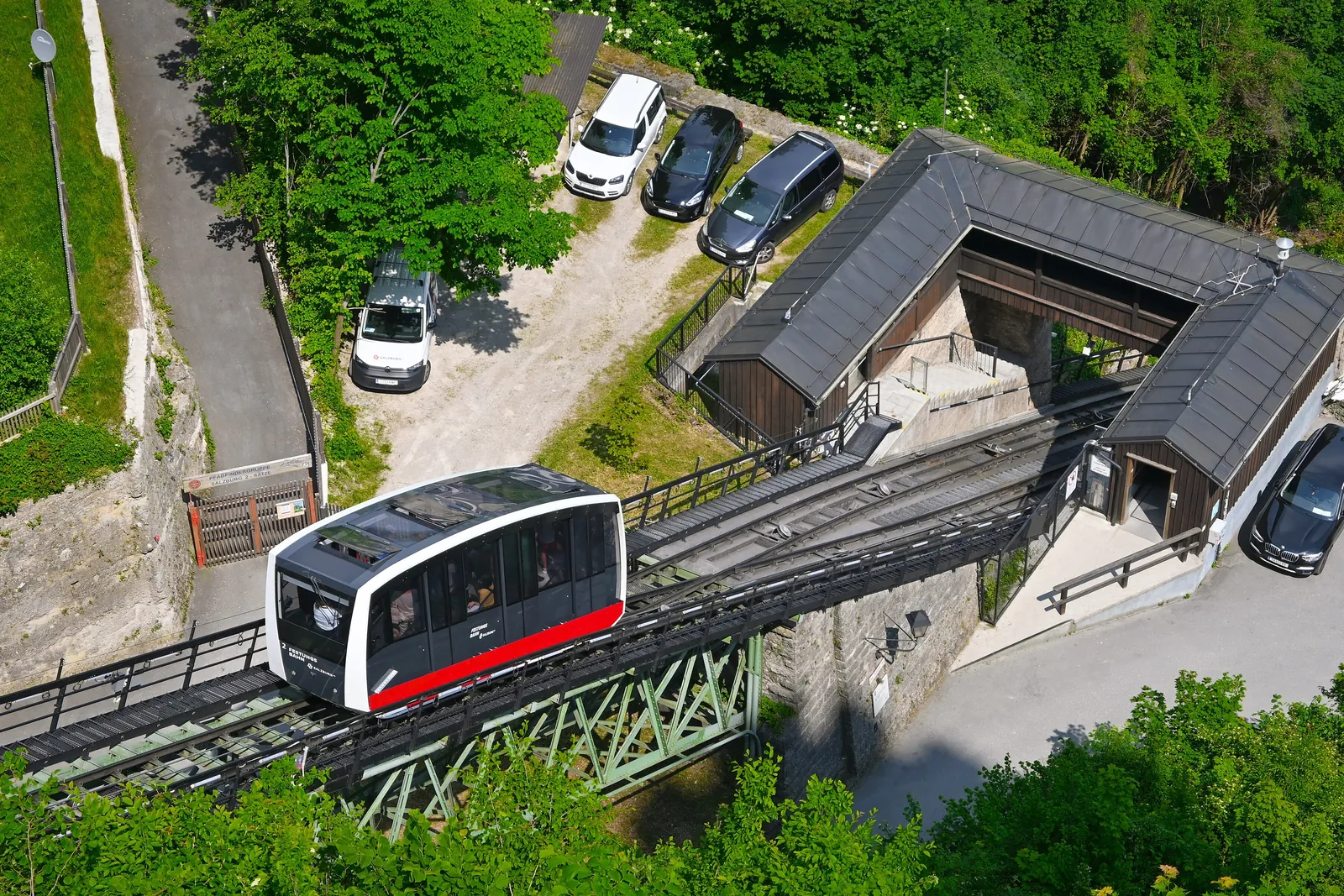 Von oben gesehen, eine Standseilbahnkabine an der Talstation der FestungsBahn zwischen Bäumen, mit dem Stationsdach und einem kleinen Parkplatz daneben
