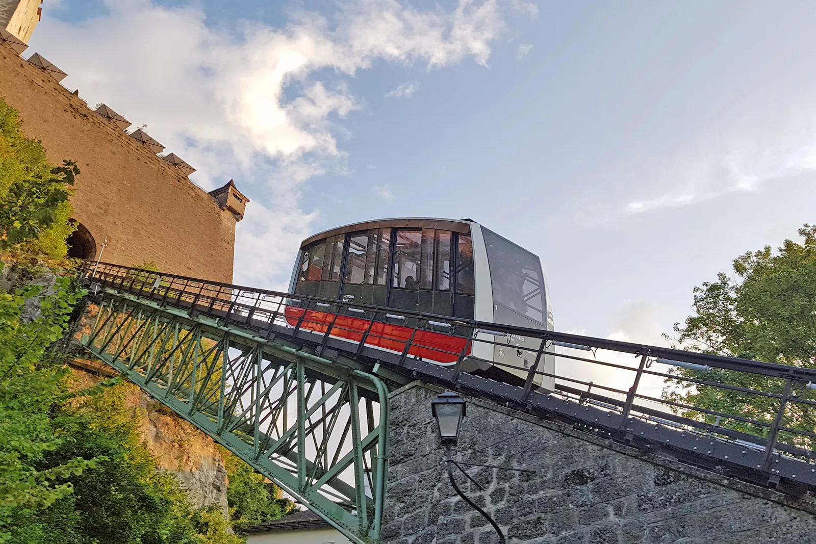 Die Standseilbahn FestungsBahn überquert eine grüne Stahlbrücke auf ihrem Weg den Hügel hinauf, mit einer Burgmauer dahinter und blauem Himmel darüber