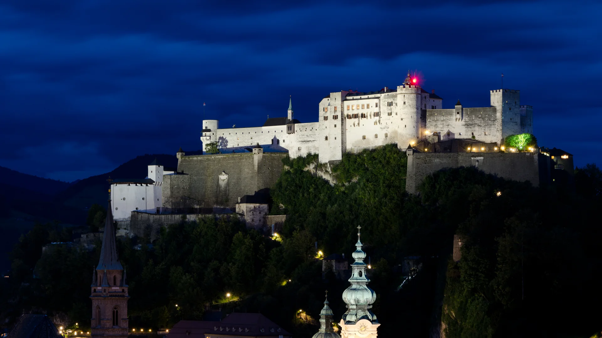 Die Festung Hohensalzburg hell erleuchtet auf ihrem Hügel vor einem tiefblauen Abendhimmel, mit den grünen Kuppeln und Türmen der Salzburger Altstadt im Vordergrund