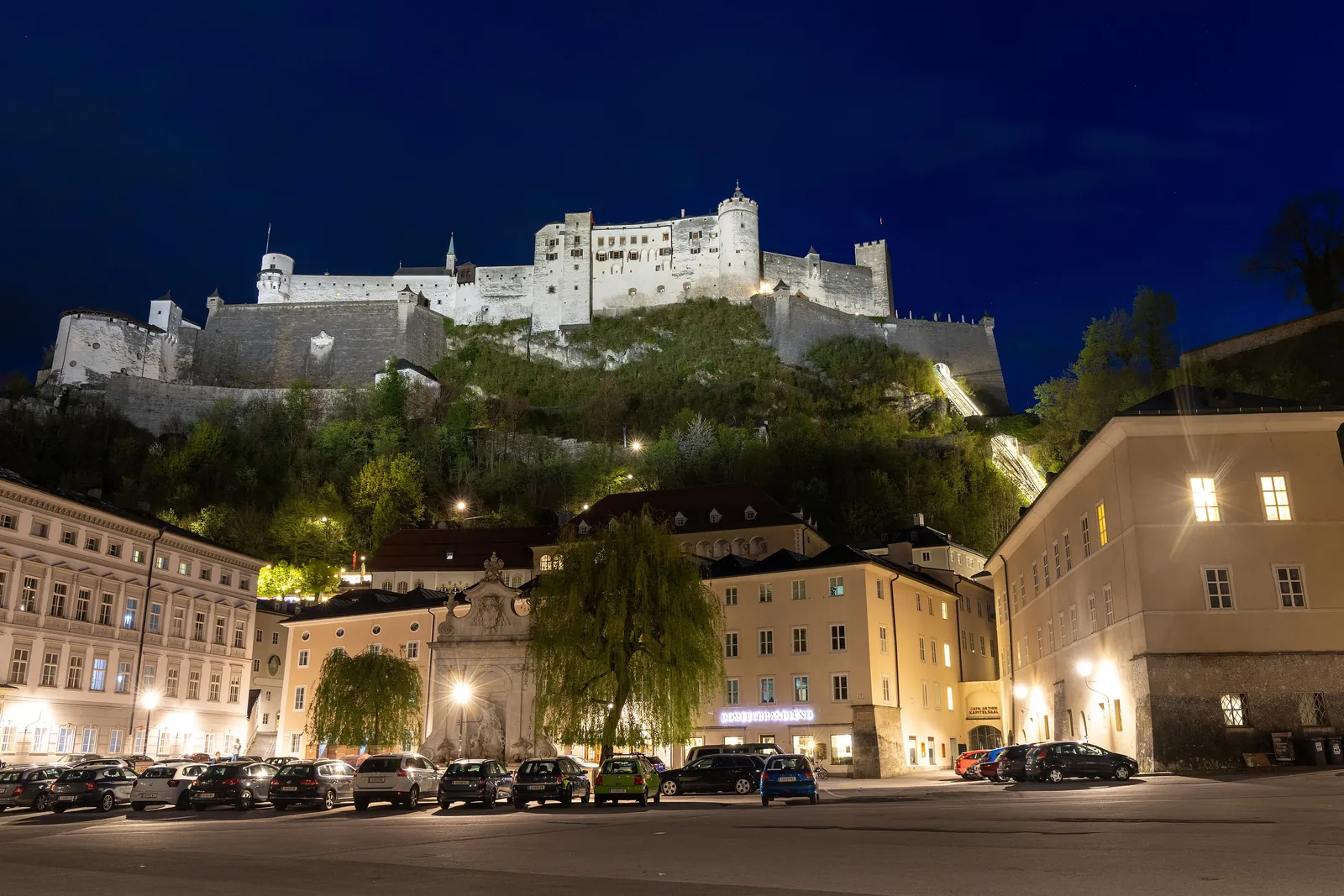 Die Festung Hohensalzburg angestrahlt über einem angestrahlten Platz in der Altstadt bei Nacht, mit geparkten Autos und einem Baum im Vordergrund