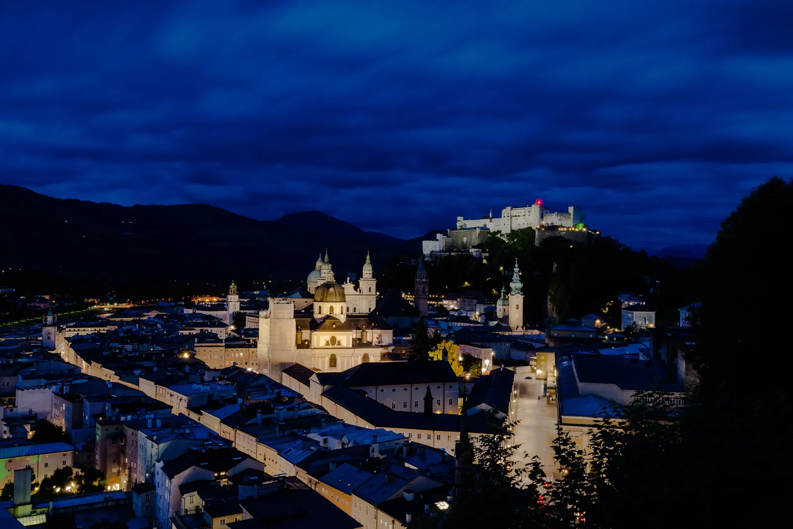 Salzburgs Altstadt in der Abenddämmerung mit beleuchteten Straßen und Kirchendomen, und die Festung leuchtet auf dem Hügel in der Ferne unter einem dunkelblauen Himmel