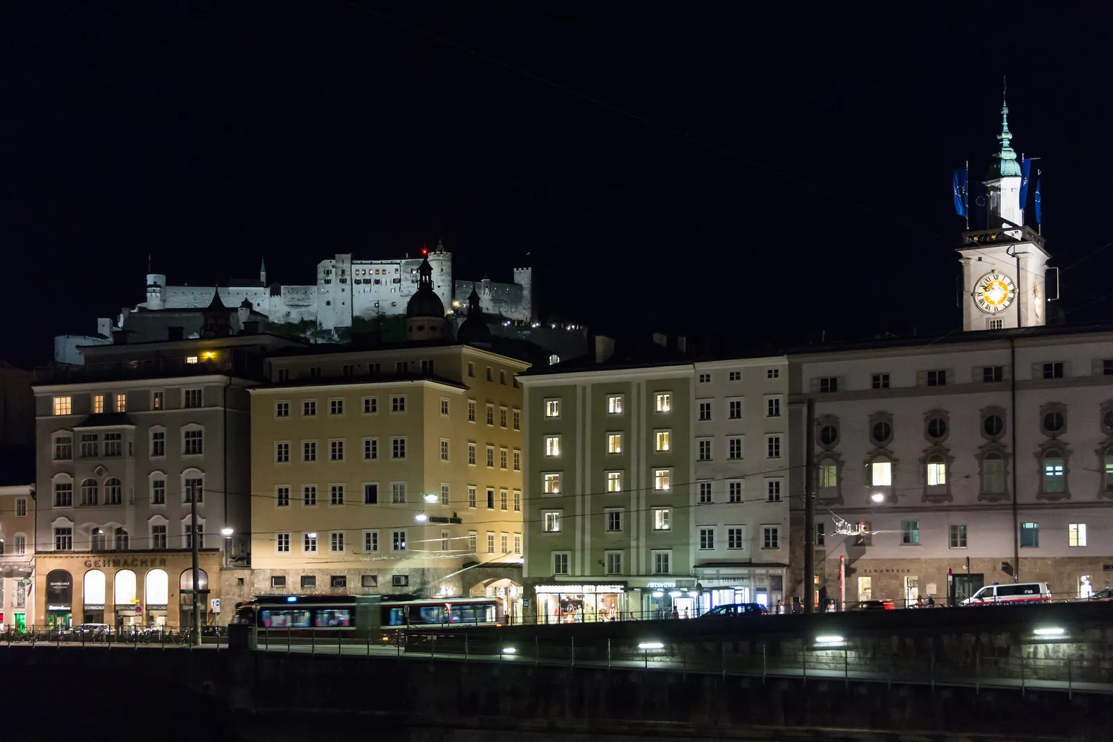 Häuser entlang des Salzachufers bei Nacht beleuchtet, mit dem Altstadt-Glockenturm rechts und der Festung, die über den Dächern leuchtet