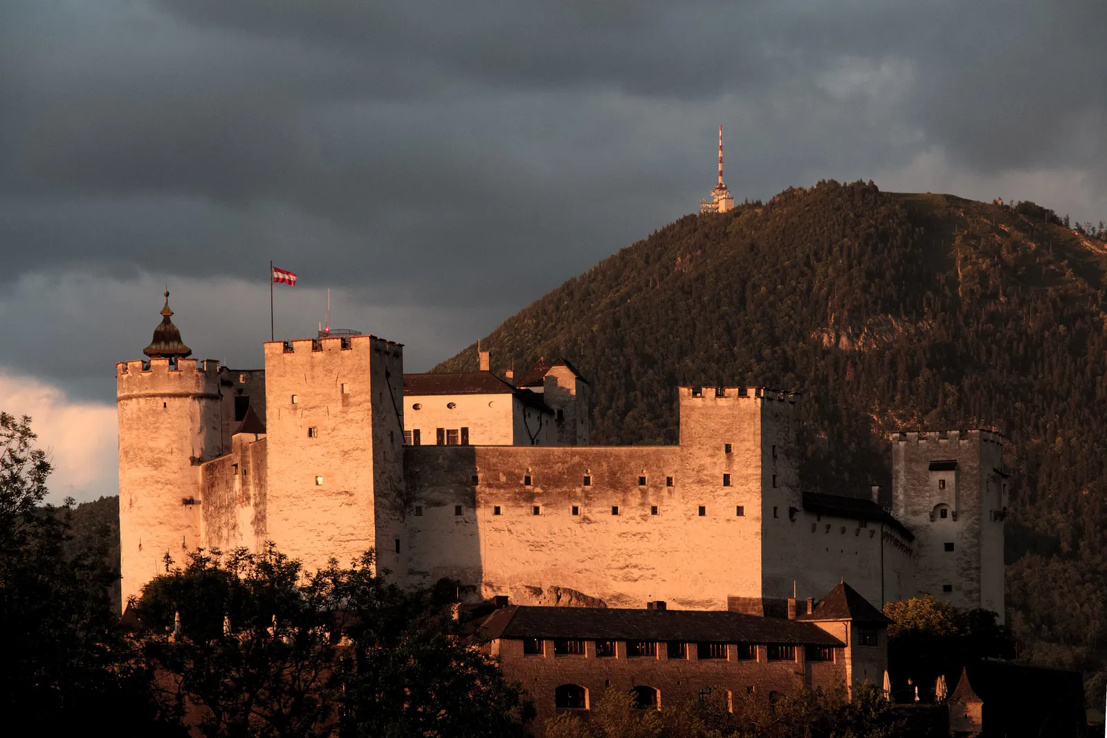 Die Türme und Mauern der Festung Hohensalzburg orange beleuchtet von der tiefen Sonne, mit dem bewaldeten Gaisberg dahinter aufragend